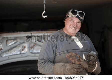 Cheerful male auto mechanic in dirty clothes in the garage holds a grinder.