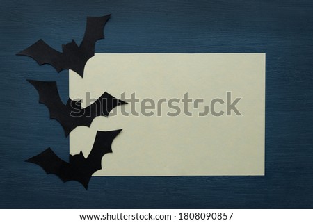 Three paper black bats and a blank sheet of light paper on a dark background with space for text. Selective focus. Copy space. Halloween greeting card