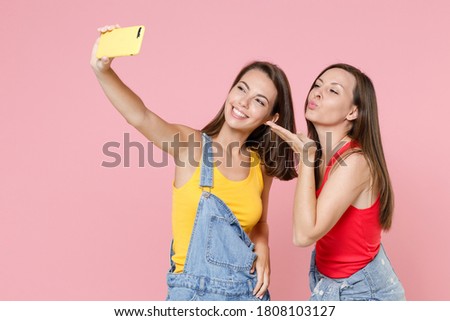 Two smiling pretty young brunette women friends 20s wearing casual denim clothes doing selfie shot on mobile phone blowing sending air kiss isolated on pastel pink colour background, studio portrait