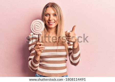Young blonde woman holding lollipop smiling happy and positive, thumb up doing excellent and approval sign 