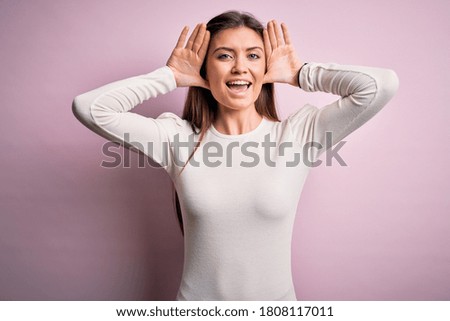 Young beautiful woman with blue eyes wearing casual white t-shirt over pink background Smiling cheerful playing peek a boo with hands showing face. Surprised and exited