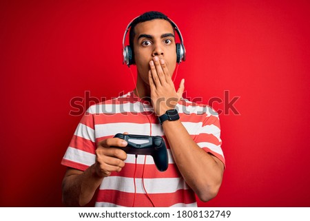 Handsome african american gamer man playing video game using jostick and headphones cover mouth with hand shocked with shame for mistake, expression of fear, scared in silence, secret concept