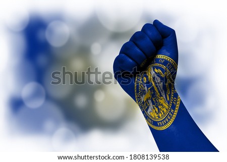 Flag of State of Nebraska painted on male fist, strength,power,concept of conflict. On a blurred background.
