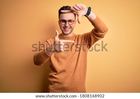 Young handsome caucasian man wearing glasses and casual winter sweater over yellow background smiling making frame with hands and fingers with happy face. Creativity and photography concept.