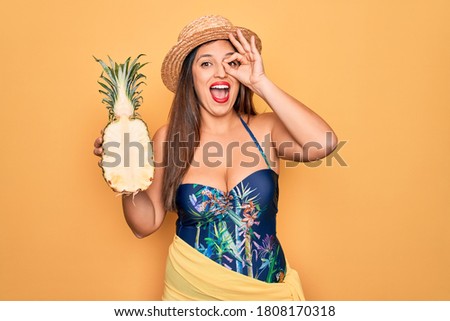 Young hispanic woman wearing summer hat and swimsuit holding tropical pineapple with happy face smiling doing ok sign with hand on eye looking through fingers