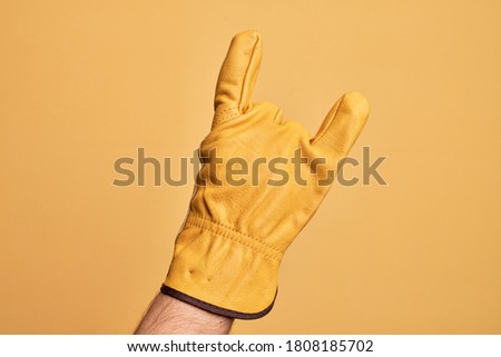 Hand of caucasian young man with gardener glove over isolated yellow background gesturing rock and roll symbol, showing obscene horns gesture