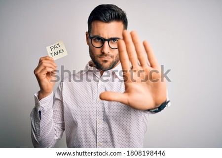 Young handsome man holding reminder paper with pay taxes message over white background with open hand doing stop sign with serious and confident expression, defense gesture