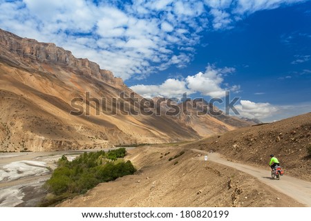 mountain bike rides along the winding dirt road in the high mountains along the mountain stream. In view of the rear. Background blue sky with clouds, Himalayas, Indian Tibet