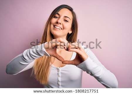 Young beautiful blonde woman with blue eyes wearing white t-shirt over pink background smiling in love doing heart symbol shape with hands. Romantic concept.