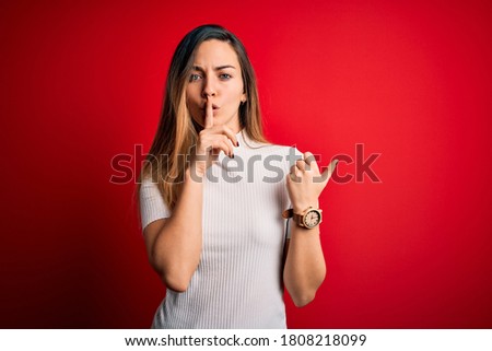 Beautiful blonde woman with blue eyes wearing casual white t-shirt over red background asking to be quiet with finger on lips pointing with hand to the side. Silence and secret concept.