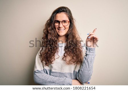 Young beautiful woman with curly hair wearing sweater and glasses over white background with a big smile on face, pointing with hand and finger to the side looking at the camera.