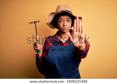 Young African American afro farmer woman with curly hair wearing apron and hat using rake with open hand doing stop sign with serious and confident expression, defense gesture