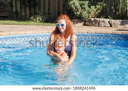 Red-haired Caucasian mother training baby son to swim in swimming pool outdoor. Baby boy in water goggles diving in water. Water family activity outdoor. Healthy active lifestyle. 