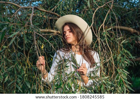 Young beautiful brunette woman dressed in a white sweater, jeans and cowboy straw hat standing close to the tree brunches with green leaves