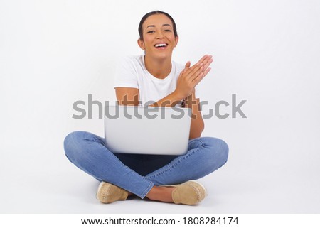 Young successful woman using computer standing against white wall, feeling happy, smiling and clapping hands, saying congratulations with an applause.