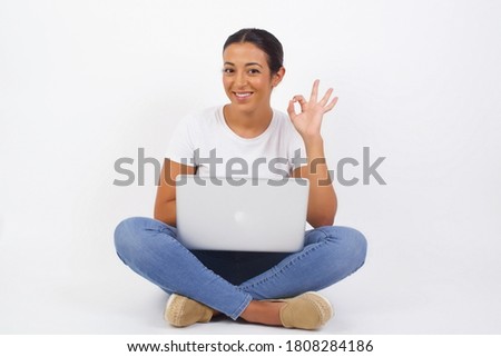 Glad attractive woman using computer shows ok sign with hand as expresses approval, has cheerful expression, being optimistic. Standing against white wall.