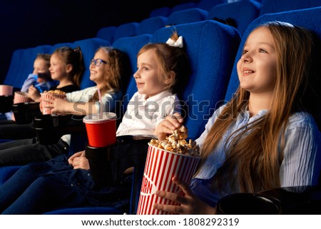 Selective focus of smiling little girl holding popcorn bucket, sitting with laughing friends in comfortable chairs in cinema. Children watching cartoon or movie, enjoying time. Entertainment concept.