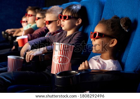 Happy african female child wearing 3d glasses, holding popcorn and watching comical movie. Cute little girl spending free time with friends in cinema and laughing. Concept of leisure, entertainment.