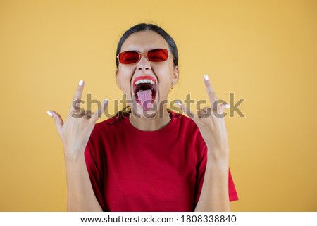 Young beautiful woman wearing red sunglasses over isolated yellow background shouting with crazy expression doing rock symbol with hands up.