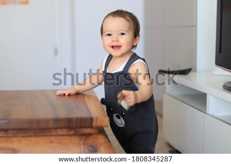Cute baby boy first step standing trying to walk with hand holding table. Mixed race Asian-German infant about 9-10 months old smiling with teething. Happy kid playing at home. Royalty-Free Stock Photo #1808345287