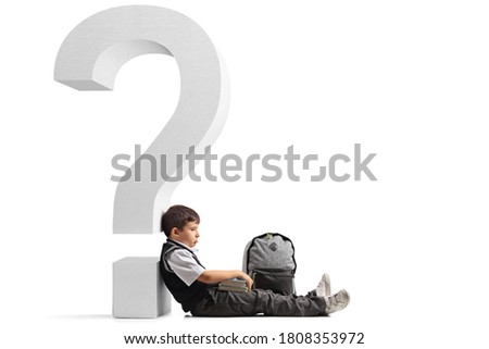 Sad schoolboy with a backpack and books seated on the floor and leaning on a question mark isolated on white background