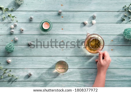 Wintertime tea, arrangement with glass teapot, glass of tea in hand on light blue mint wooden background. Christmas decorations, disco balls, baubles, toys, tea candle and eucalyptus.