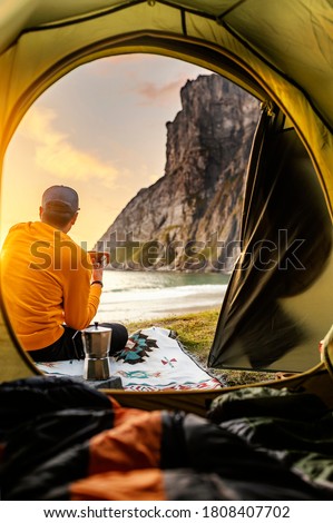 Camping with tent in Lofoten islands, Northern Norway, Kvalvika beach, during sunset. Person sitting in front of a tent holding a hot drink mug. Royalty-Free Stock Photo #1808407702