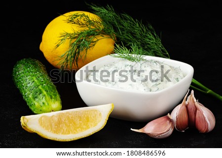 Bowl of fresh tzatziki sauce with cucumber, garlic, dill and lemon on black background. Yogurt dip. Greek cuisine. Closeup.