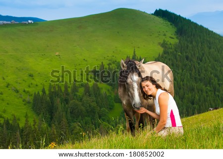 happy girl sitting on a green flower meadow near a wild horse and her calf. background of green hills