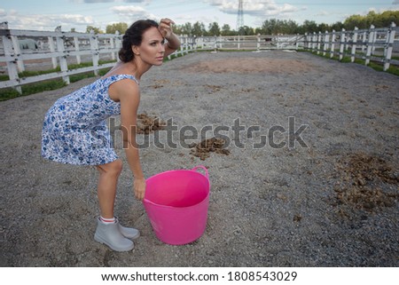 A woman in a horse paddock removes horse manure, pretends to be a tired man, a comical photo. Agriculture concept