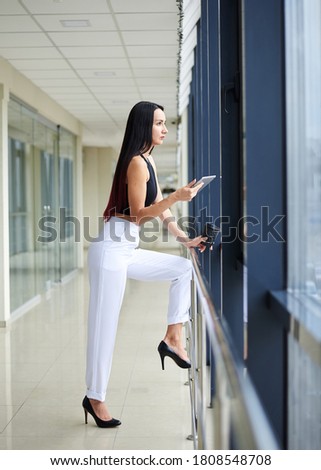 Young brunette woman, wearing white pants and black top, holding phone, standing by the window in light office building. Office worker on break.