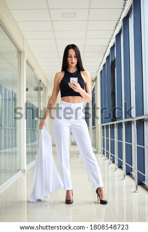 Young brunette woman, wearing white pants and black top, holding jacket in hand, taking selfie picture with cell phone in light passageway, posing for social media. Businesswoman on lunch break.