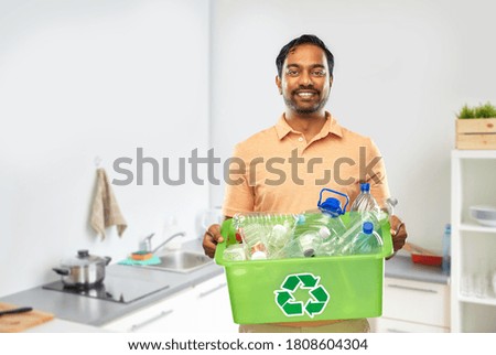 recycling, waste sorting and sustainability concept - smiling young indian man in striped t-shirt holding box with plastic bottles over home kitchen background