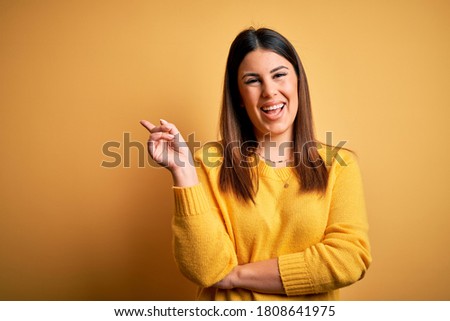 Young beautiful woman wearing casual sweater over yellow isolated background with a big smile on face, pointing with hand and finger to the side looking at the camera.