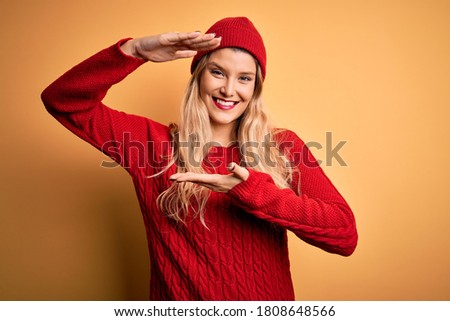 Young beautiful blonde woman wearing casual sweater and wool cap over white background gesturing with hands showing big and large size sign, measure symbol. Smiling looking at the camera. Measuring.