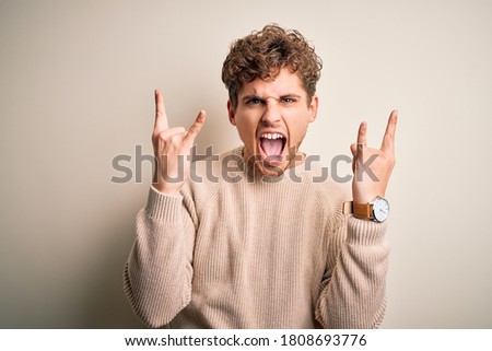 Young blond handsome man with curly hair wearing casual sweater over white background shouting with crazy expression doing rock symbol with hands up. Music star. Heavy concept.