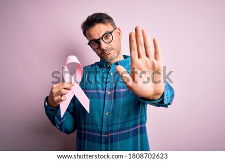 Young handsome man holding pink cancer ribbon symbol over isolated background with open hand doing stop sign with serious and confident expression, defense gesture