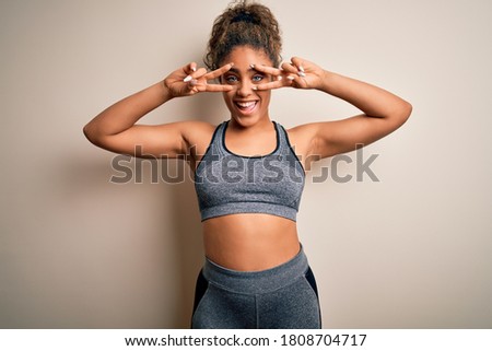 Young african american sportswoman doing sport wearing sportswear over white background Doing peace symbol with fingers over face, smiling cheerful showing victory