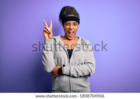 Young african american sportswoman doing sport wearing sportswear over purple background smiling with happy face winking at the camera doing victory sign. Number two.