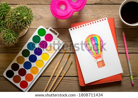 palette with watercolors, brushes, cactus, cup of coffee, pencil, colorful balloon drawing on white paper on wooden table Top view Flat lay Сreation, hobby and education concept Stay home