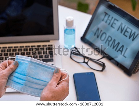 Woman's hand holding a surgical mask against covid-19 infection- laptop computer and tablet with message - technological devices on the table