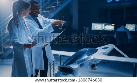 Two Aerospace Engineers Work On Unmanned Aerial Vehicle / Drone Prototype. Aviation Scientists Talking, using Blueprints. Industrial Laboratory with Commercial Aerial Surveillance Aircraft Royalty-Free Stock Photo #1808746030