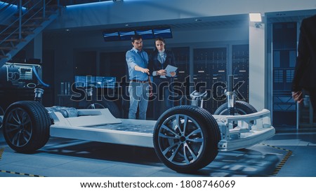 Auto Industry Design Facility: Male Chief Engineer Shows Car Prototype to Female Car Designer. Electric Vehicle Platform Chassis Concept with Wheels, Engine and Battery.