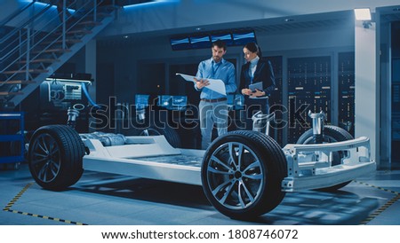 Auto Industry Design Facility: Male Chief Engineer Shows Car Blueprints Female Software Design and Integration Engineer. Electric Vehicle Platform Chassis Concept Has Wheels, Engine and Battery