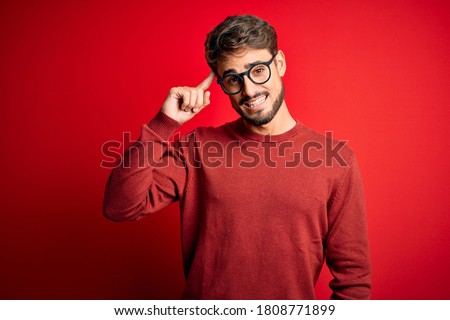 Young handsome man with beard wearing glasses and sweater standing over red background Smiling pointing to head with one finger, great idea or thought, good memory