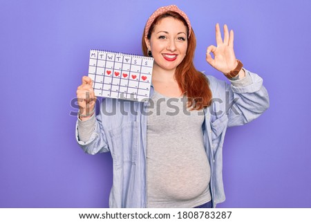 Young beautiful redhead pregnant woman expecting baby holding period calendar doing ok sign with fingers, excellent symbol