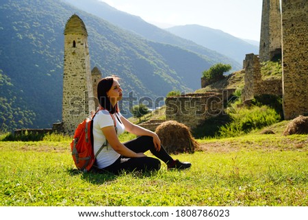 Carefree traveling woman relaxing on green lawn. Side view of serene female tourist with backpack sitting on green hill and enjoying sunny weather on background of mountainous landscape