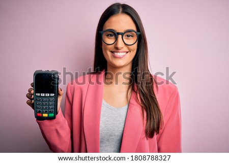 Young beautiful brunette woman holding dataphone paying with credit card to finance with a happy face standing and smiling with a confident smile showing teeth