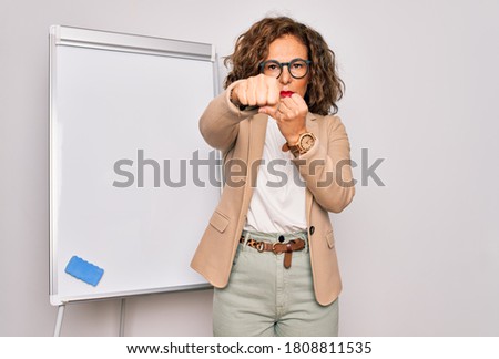 Middle age senior business woman standing on seminar presentation by magnectic blackboard Punching fist to fight, aggressive and angry attack, threat and violence