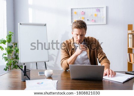 thoughtful businessman looking at laptop while sitting at workplace near notebook, documents and coffee cup